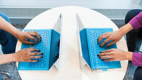 Two people typing on laptops on a small table. 