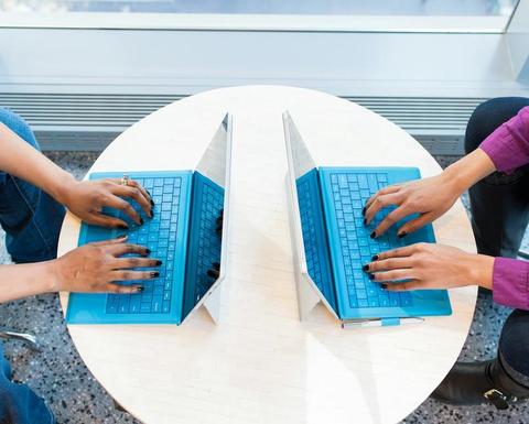 Two people typing on laptops on a small table. 